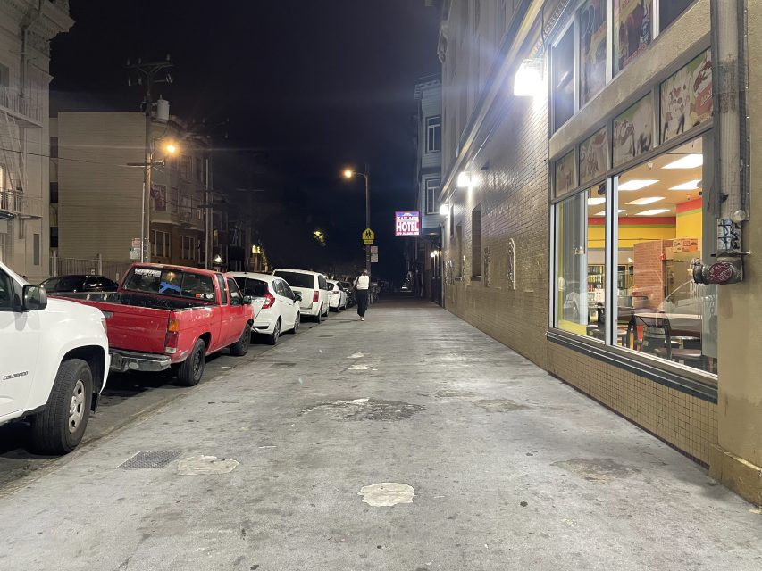 A city street at night with parked cars on the left, a lit restaurant on the right, and a person walking in the distance under streetlights.