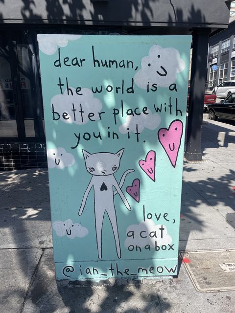 A painted utility box features a cartoon cat, pink hearts, and clouds with text that reads: "dear human, the world is a better place with you in it. love, a cat on a box.
