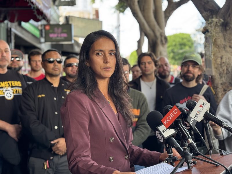 A woman in a purple blazer speaks at a podium with multiple microphones, while a group of people stand behind her outdoors.