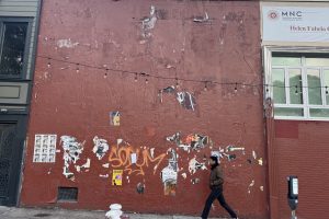A person walks past a red wall with peeling posters, graffiti, and string lights above; part of a building and a hydrant are visible.