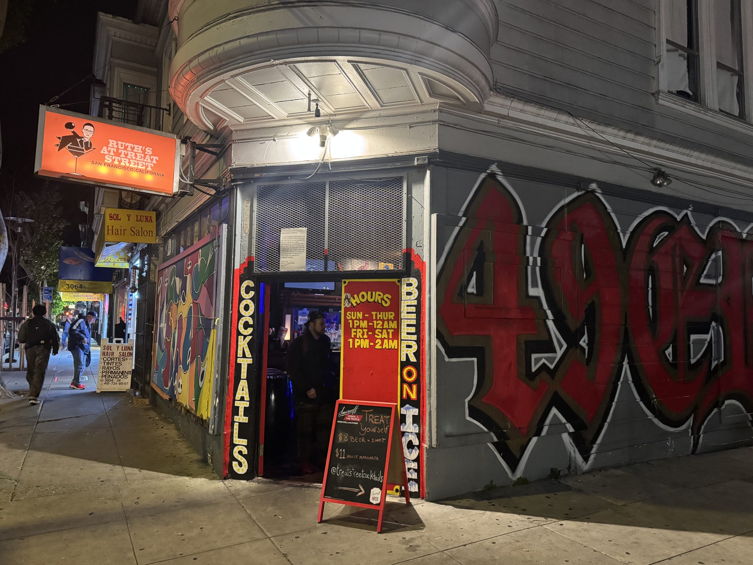 A bar entrance with a "COCKTAILS" sign, business hours posted, and a large "49ERS" mural on the wall outside at night. People are visible on the sidewalk.