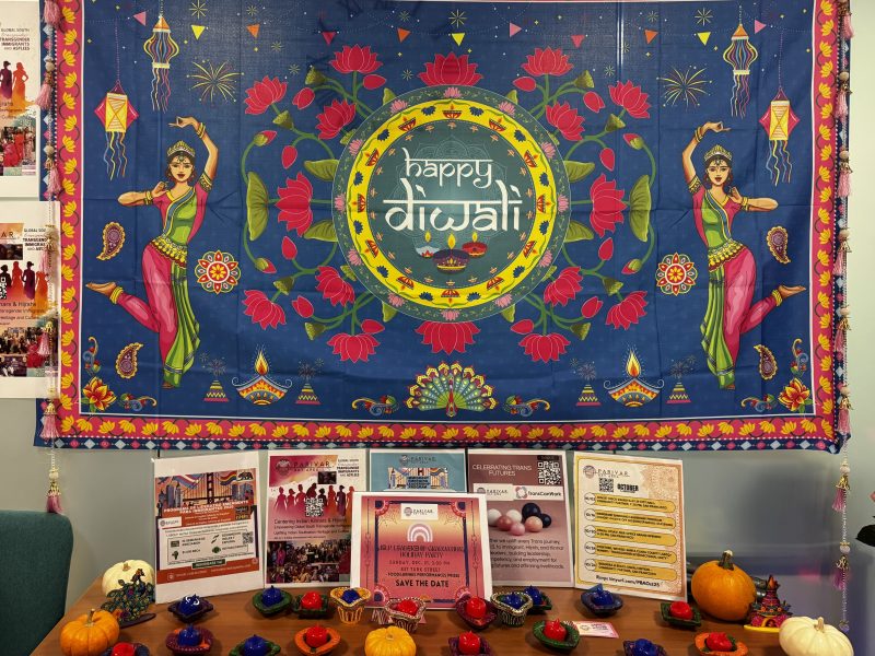A colorful "Happy Diwali" banner with floral and dancer designs, including transgender representation, hangs above a table decorated with small pumpkins, candles, and informational flyers.