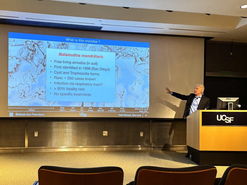 A presenter at a UCSF lecture points to a slide about Balamuthia mandrillaris, detailing its characteristics, rarity, high fatality rate, and lack of specific treatments.