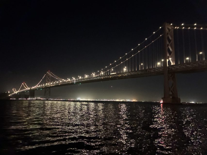 Wide view of a suspension bridge illuminated at night, with lights reflecting on the water and a city skyline in the background.