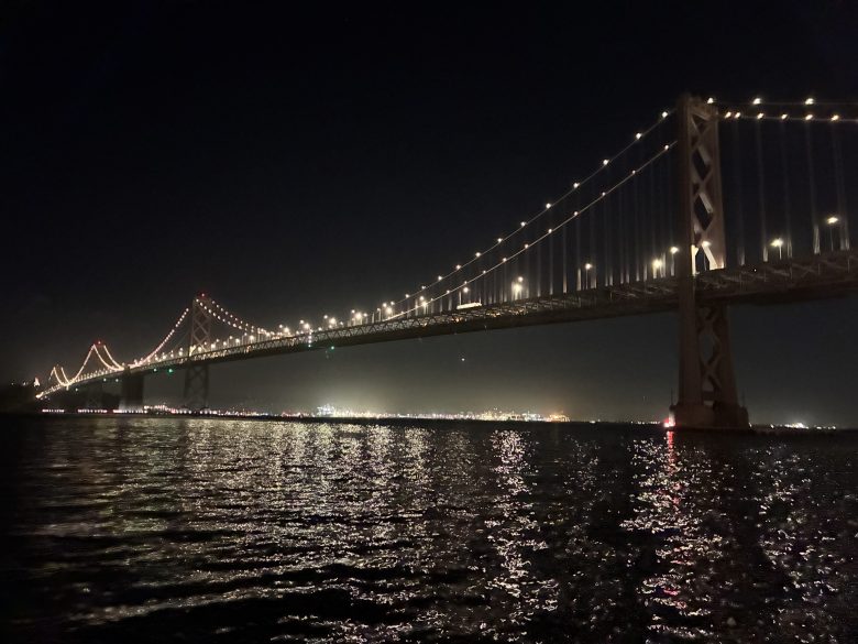 A large suspension bridge is illuminated at night, reflecting lights on the water below, with a cityscape visible in the background.