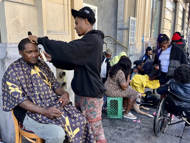 A man gives a haircut to another man seated on a sidewalk, while several people gather nearby, some sitting and one in a wheelchair.