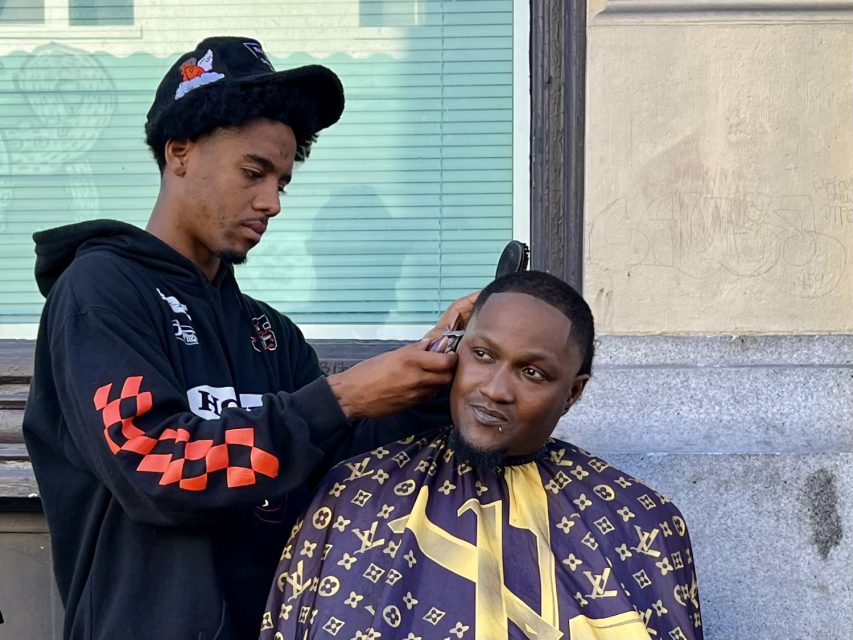 A barber gives a man a haircut outdoors in front of a building, with the client wearing a patterned cape and the barber using electric clippers.