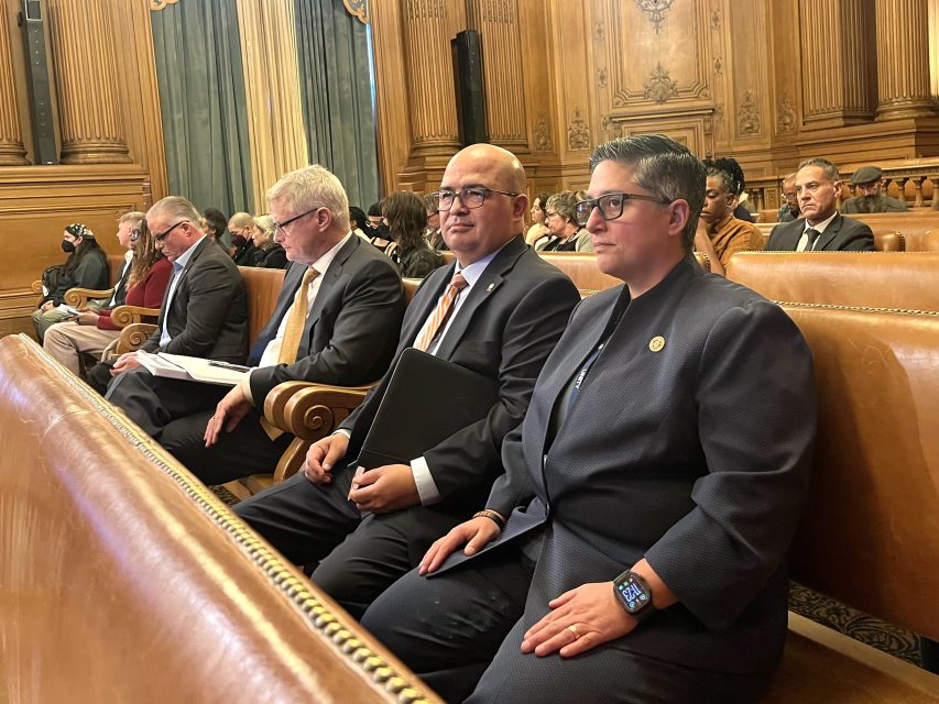 People in formal attire sit in wooden benches inside a government or courtroom setting, with others seated in the background.