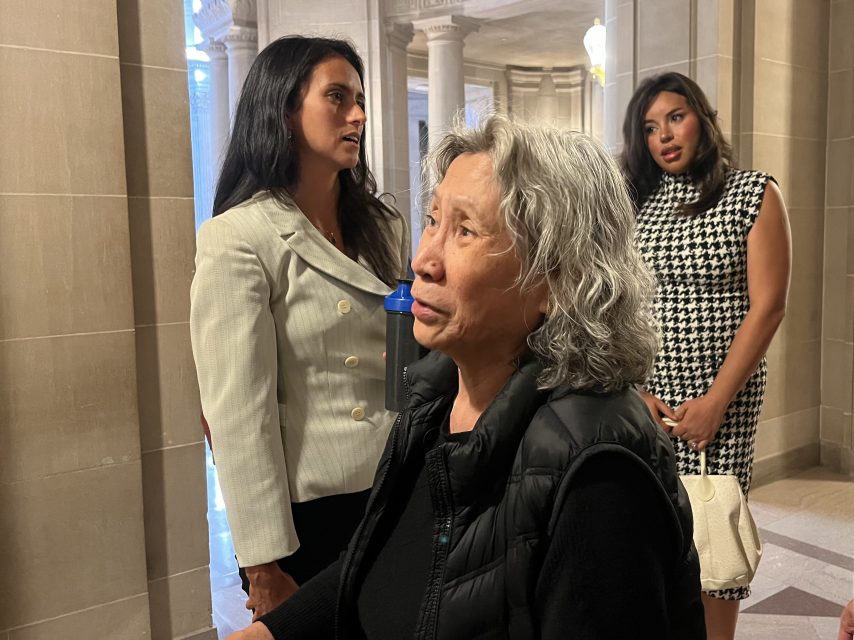 Three women stand indoors in a marble hallway, two in business attire and one in a black vest, with architectural columns visible in the background.