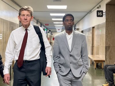 Two men in business attire walk down a hallway with beige walls and benches; one carries a bag over his shoulder.
