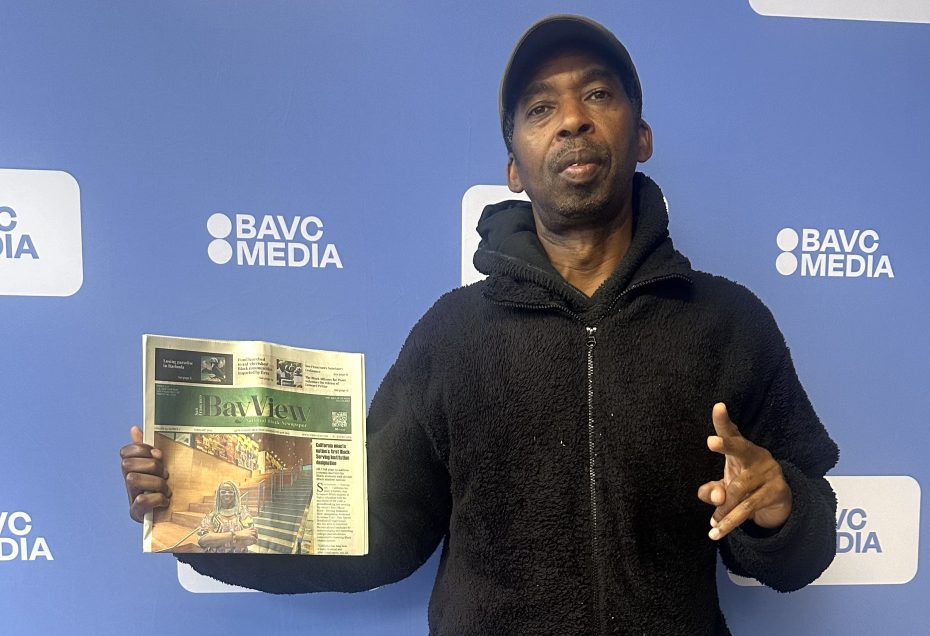 A man stands in front of a blue BAVC Media backdrop, holding a newspaper in his left hand and gesturing with his right hand.