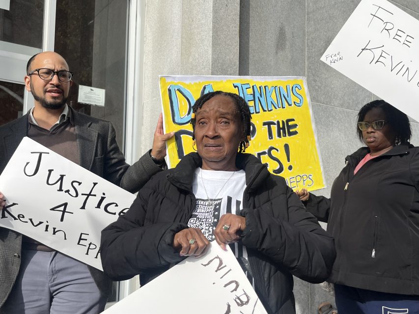 Three people stand outside holding protest signs, including ones reading "Justice 4 Kevin Epps" and "DA Jenkins DROP THE CHARGES!.