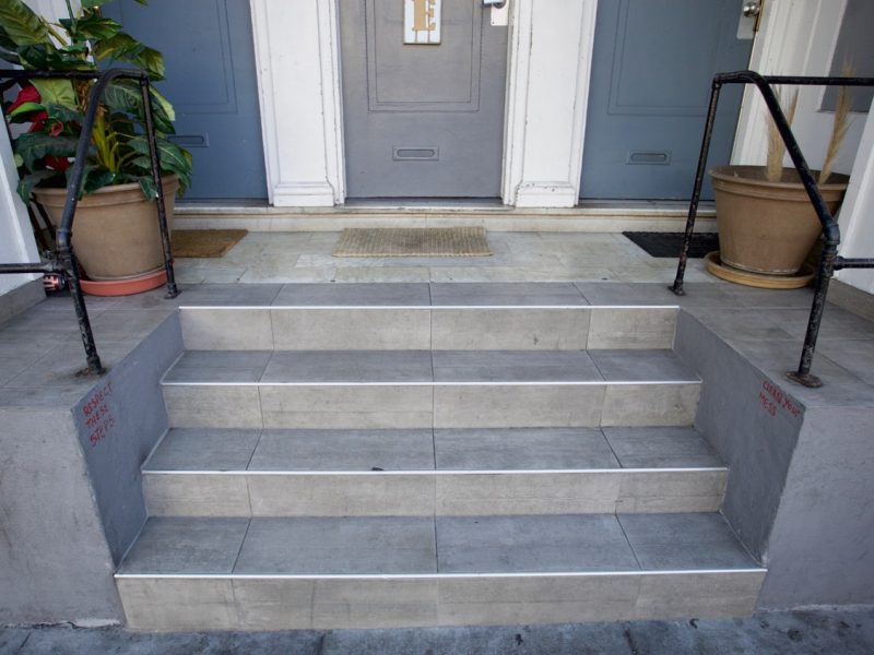Four gray tiled steps with metal edges lead up to two doors, flanked by handrails and potted plants on either side.