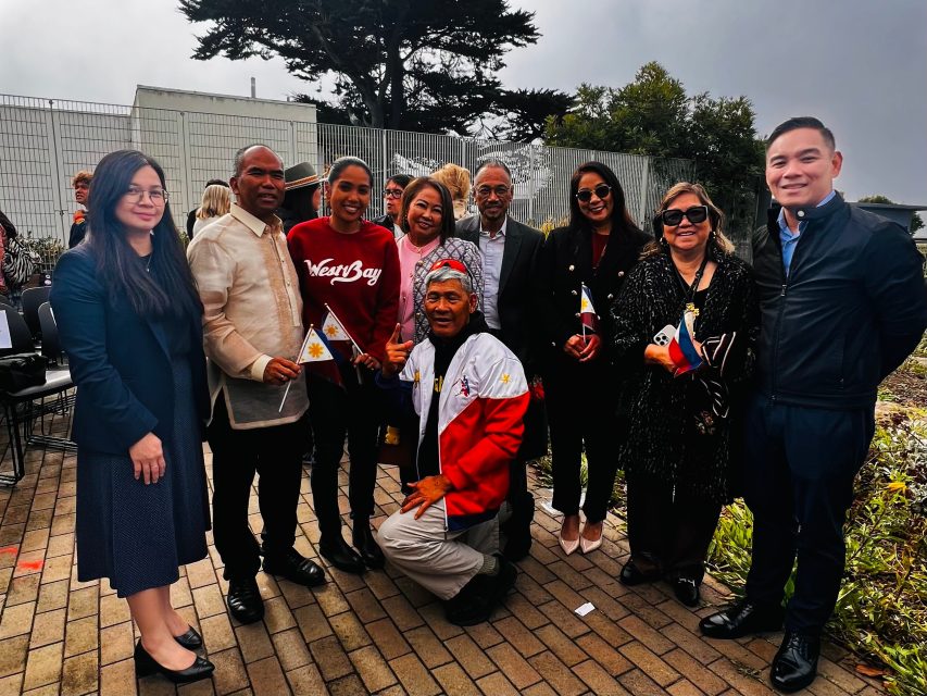 A group of people pose outdoors, some holding Philippine flags. They are smiling and dressed in a mix of casual and formal attire. Trees and a metal fence are visible in the background.