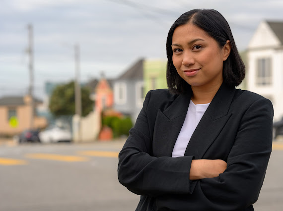 A woman in a black blazer stands with arms crossed outdoors on a street, with houses and a cloudy sky in the background.