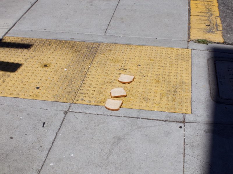 Three slices of white bread are lying on a yellow textured sidewalk curb ramp near a street corner.