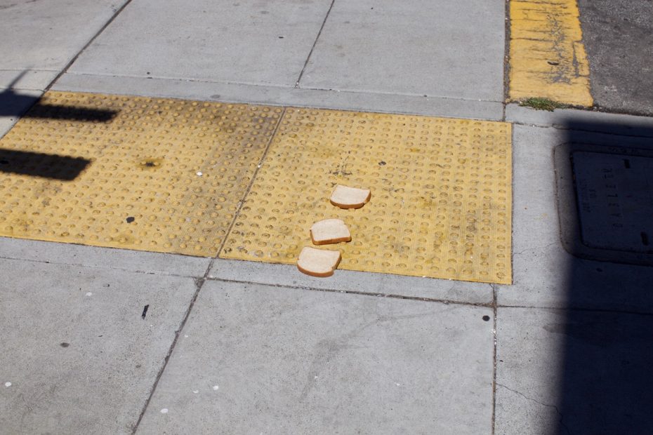Three slices of white bread are lying on a yellow textured section of a sidewalk next to a paved street.
