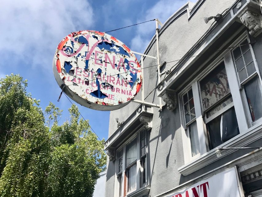 A weathered, partially faded restaurant sign hangs above the entrance of a gray building with decorative molding and windows, under a partly cloudy sky.
