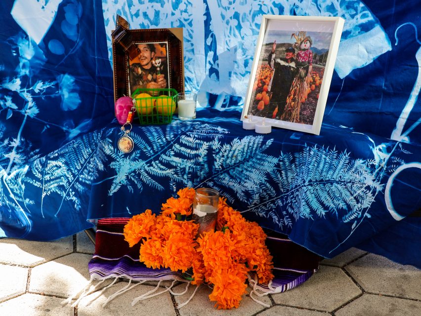 A Día de los Muertos altar with framed photos, marigold flowers, a painted skull, candles, fruit, and a pocket watch on a blue patterned cloth.