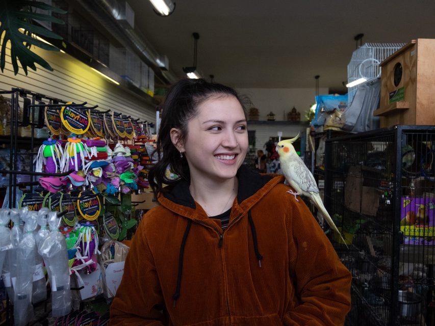 A woman in a brown jacket stands inside a pet store, smiling with a cockatiel perched on her shoulder. Colorful pet supplies are displayed on shelves in the background.