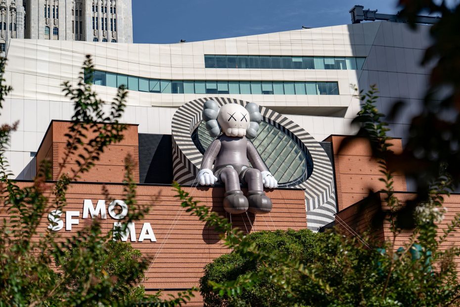 A large gray cartoon-like sculpture with X eyes sits on the roof of the SF MoMA building, surrounded by modern architecture and trees.
