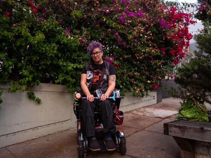 A person with purple hair and tattoos sits in a motorized wheelchair on a sidewalk, with flowering bushes in the background.