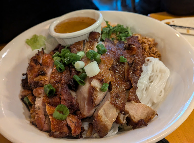 A bowl of grilled chicken slices on rice vermicelli noodles, garnished with green onions and herbs, served with a side of dipping sauce.