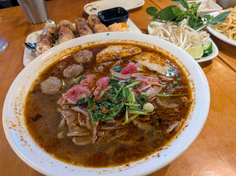 A bowl of Vietnamese spicy beef noodle soup (bun bo hue) with herbs, meat, and noodles, served alongside fresh vegetables and spring rolls on a wooden table.