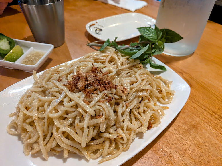 A plate of plain noodles topped with fried shallots, garnished with a sprig of basil, sits on a wooden table with a side of cucumbers, sauce, and a drink.