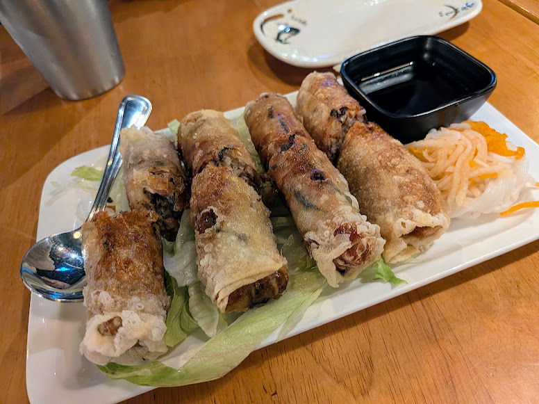 A plate of six fried spring rolls on lettuce, served with pickled vegetables, a dipping sauce, and a metal spoon on a wooden table.