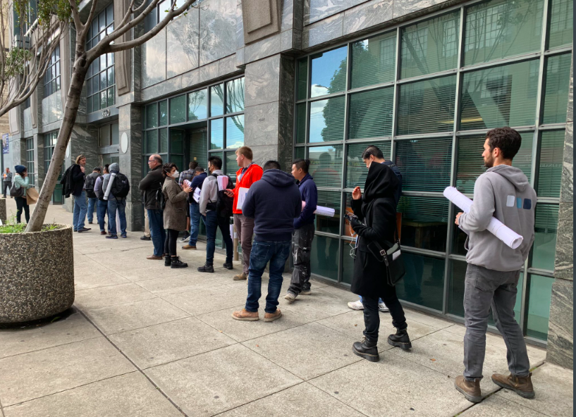 A group of people stand in line outside a building with large windows on a city sidewalk, some holding rolled-up papers.