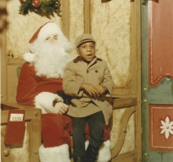 A young child in a tan coat sits on Santa Claus's lap in a festive holiday setting with decorations in the background.