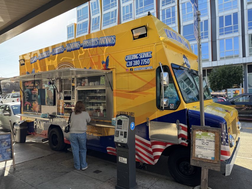 A person stands in front of a yellow and blue food truck with a service window open, parked on a city street near a sidewalk menu board.