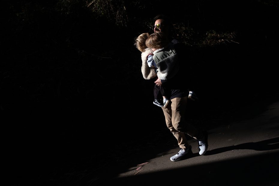 An adult wearing sunglasses carries a child while walking through a shadowed area with sunlight highlighting them.