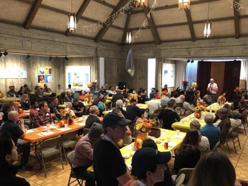 A group of people seated at tables decorated with sunflowers listens to a speaker at the front of a large hall with art on the walls.