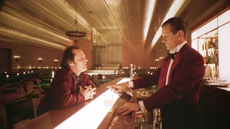 A man sits at a bar counter smiling at a bartender in a red jacket who is pouring a drink. The setting is dimly lit with art deco decor.