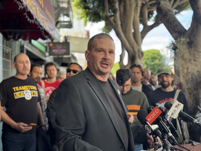 A man speaks at a podium with several microphones in front of him, while a group of people stands behind him on a city sidewalk.