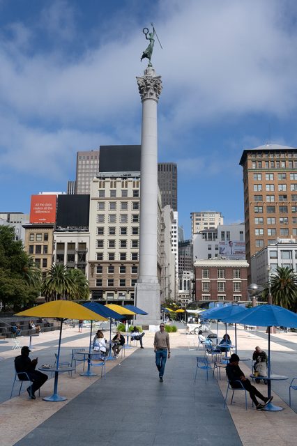 People sit and walk in an outdoor plaza with blue and yellow umbrellas, featuring a tall monument topped with a statue in the center, surrounded by city buildings.