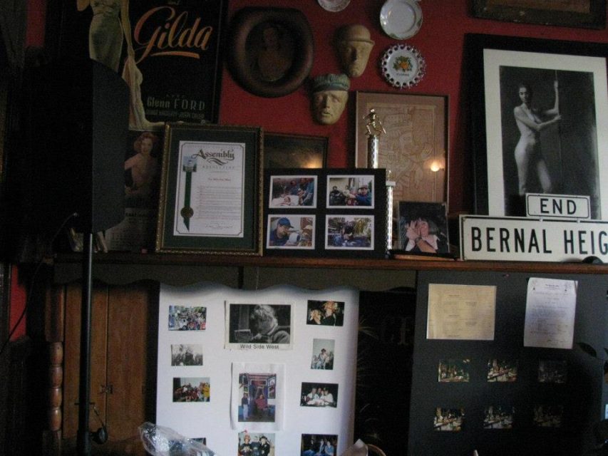 A wall filled with framed photos, posters, documents, and memorabilia, including a street sign, a trophy, and various images of people, in a dimly lit room.
