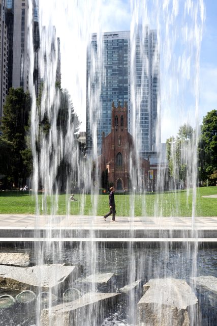 A person walks past a fountain with water jets in a city park; a historic church and modern skyscrapers are visible in the background.