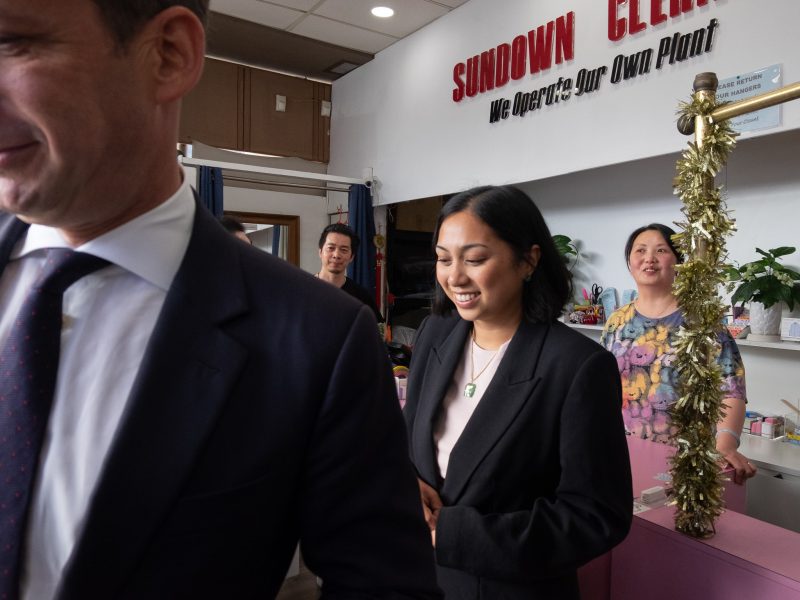 Several people stand inside a shop with a "SUNDOWN CLEANERS" sign, counter space, and festive decorations visible, while some individuals interact and others observe.
