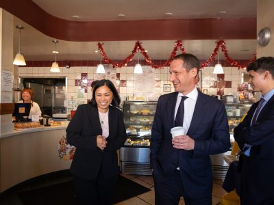 Three people in business attire talk and smile inside a bakery decorated with red garlands; a worker stands behind the counter in the background.