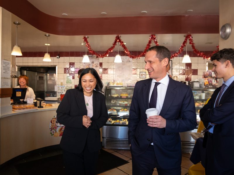 Three people in business attire talk and smile inside a bakery decorated with red garlands; a worker stands behind the counter in the background.