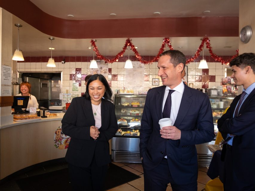 Three people in business attire talk and smile inside a bakery decorated with red garlands; a worker stands behind the counter in the background.