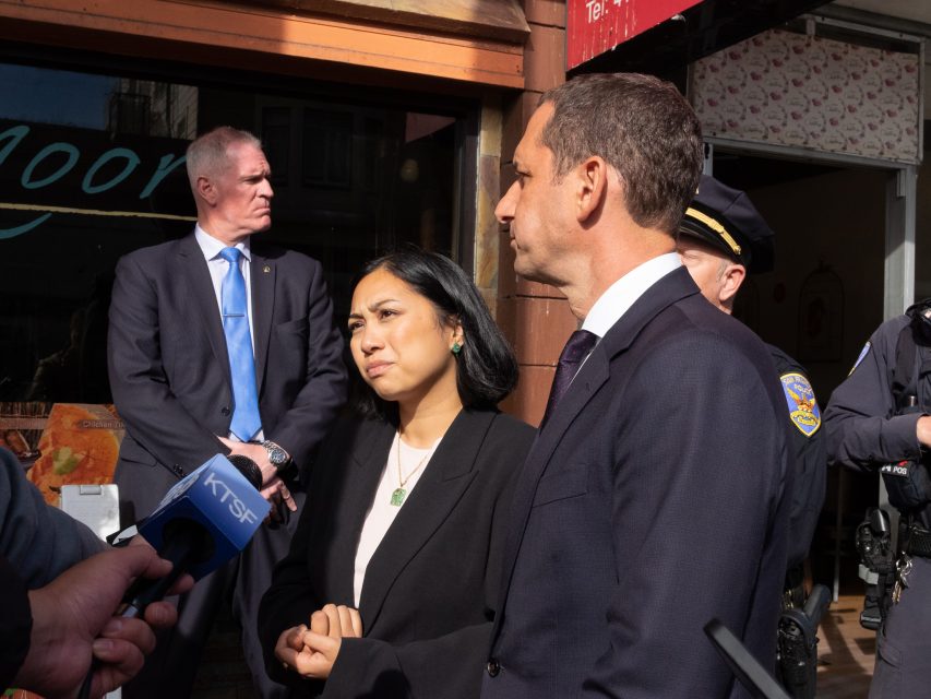 A woman and a man in business attire speak to the press outdoors, surrounded by police officers and reporters holding microphones.