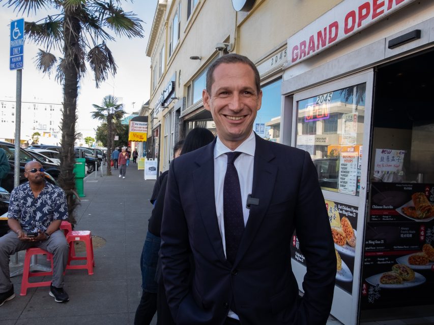 A man in a suit stands and smiles on a busy sidewalk near a restaurant with a "Grand Opening" sign; people sit and walk nearby.