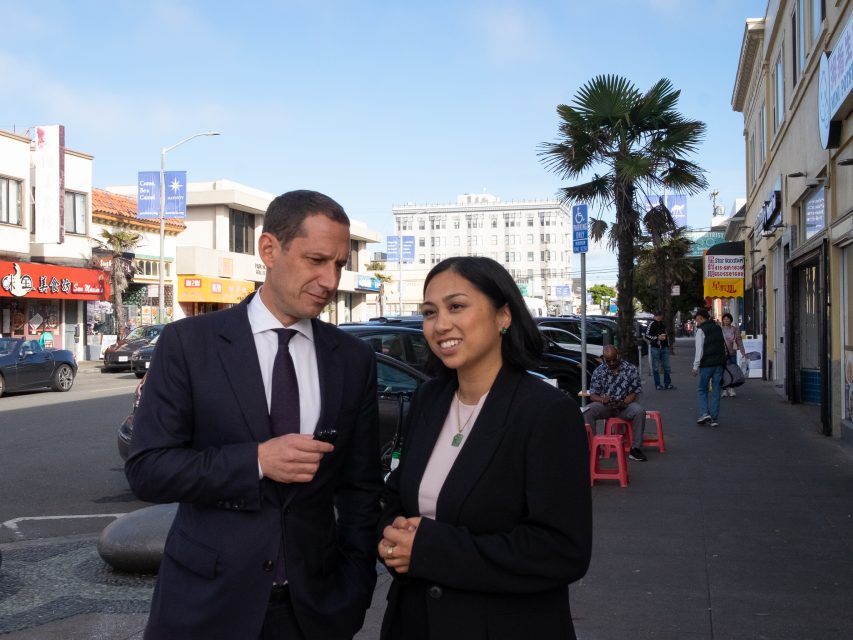 Two people in business attire stand and talk on a busy city sidewalk lined with shops, parked cars, and pedestrians in the background.