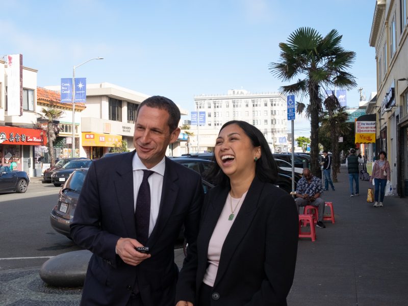 A man and a woman in business attire stand on a city sidewalk, smiling and laughing, with shops, parked cars, and pedestrians in the background.