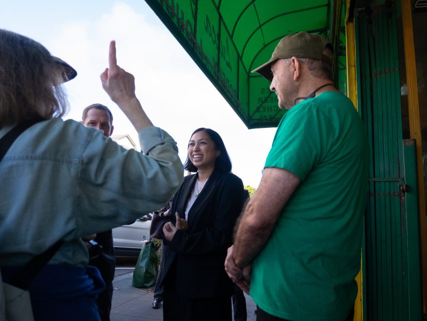 Four people stand and talk on a sidewalk under a green awning; one person gestures with their hand while another smiles.