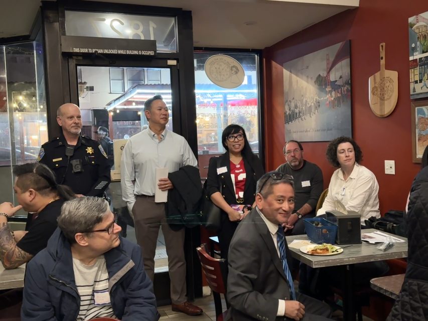 A group of people, including a police officer and professionals, are gathered inside a restaurant, some seated and some standing, listening attentively.